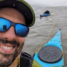 Dan Blumberg in a life jacket and cap, grinning in the rain on the Hudson, towing a second kayak behind his own